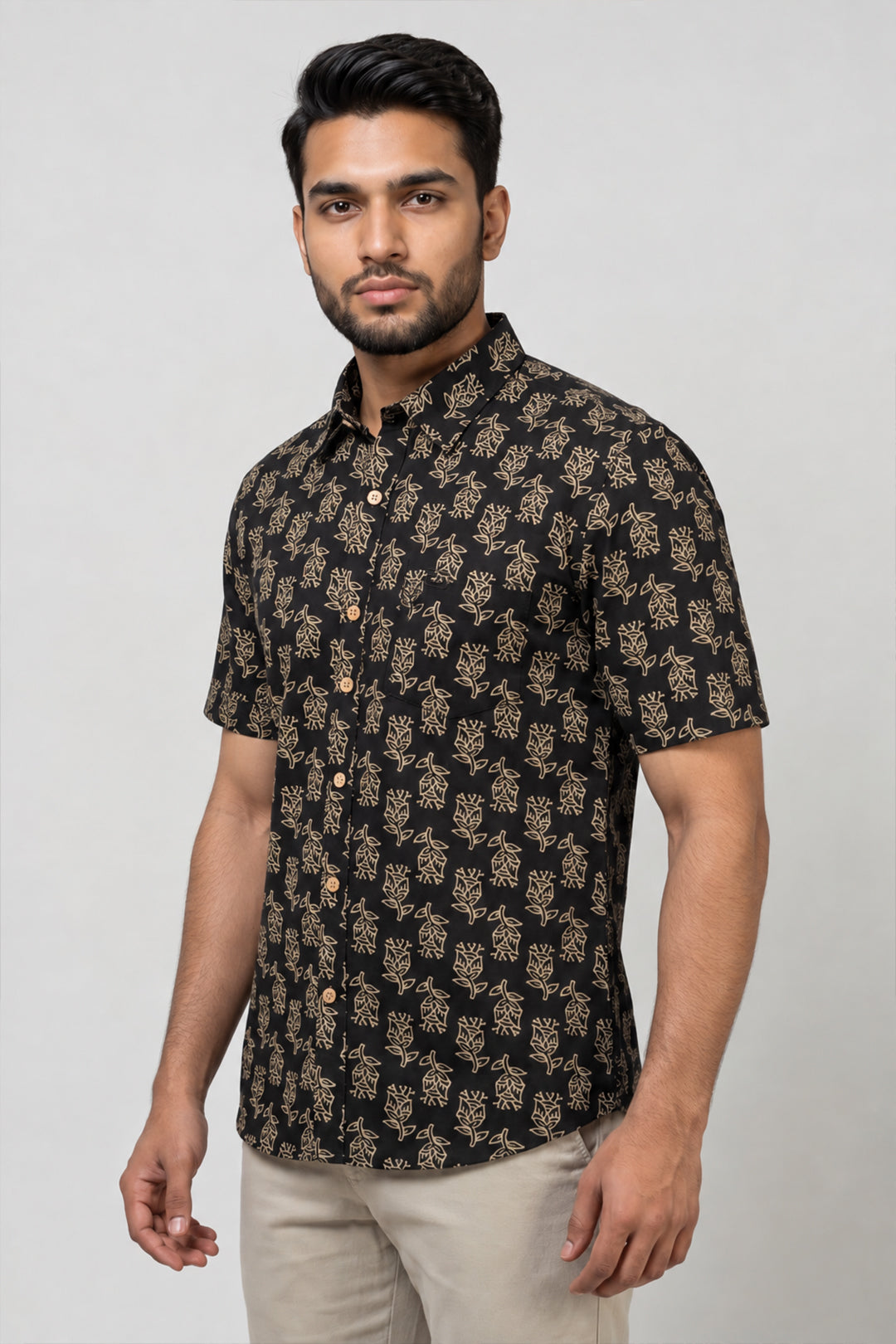 A man wearing a black short-sleeve button-up shirt with a beige floral block print pattern, paired with beige trousers, standing against a light gray studio background.