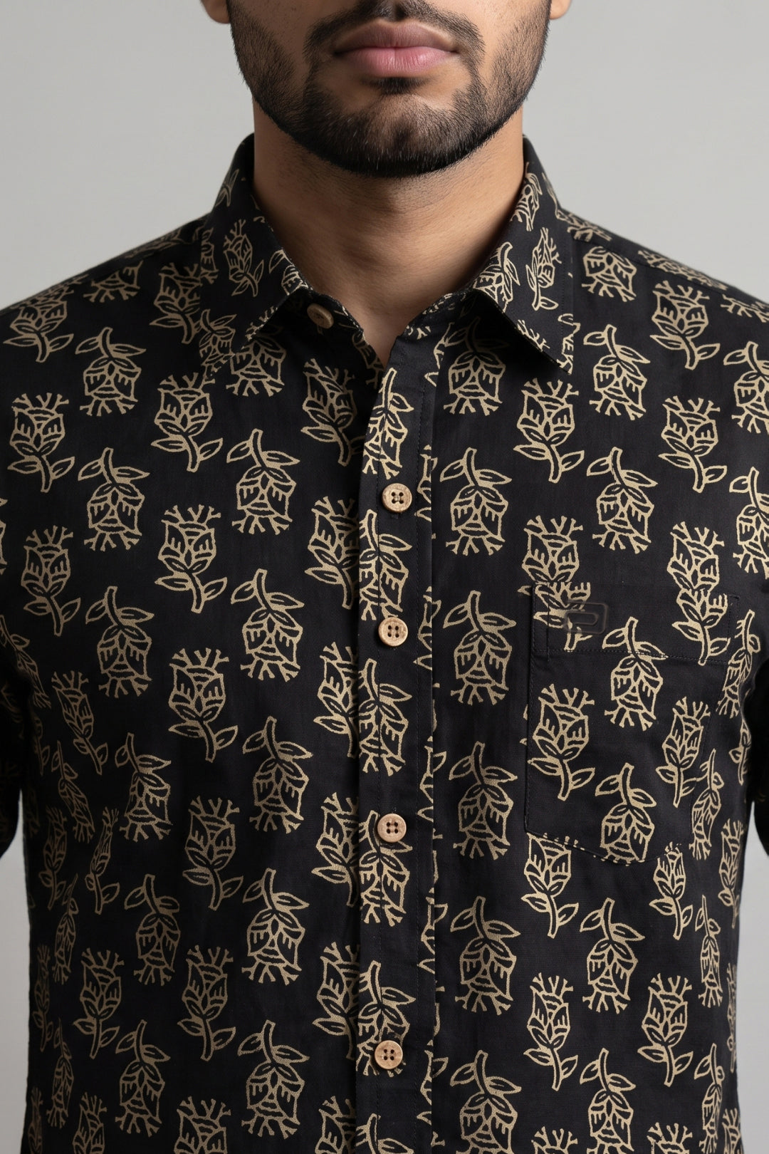 A close-up of a man wearing a black button-up shirt with a beige floral and leaf pattern, featuring brown buttons.