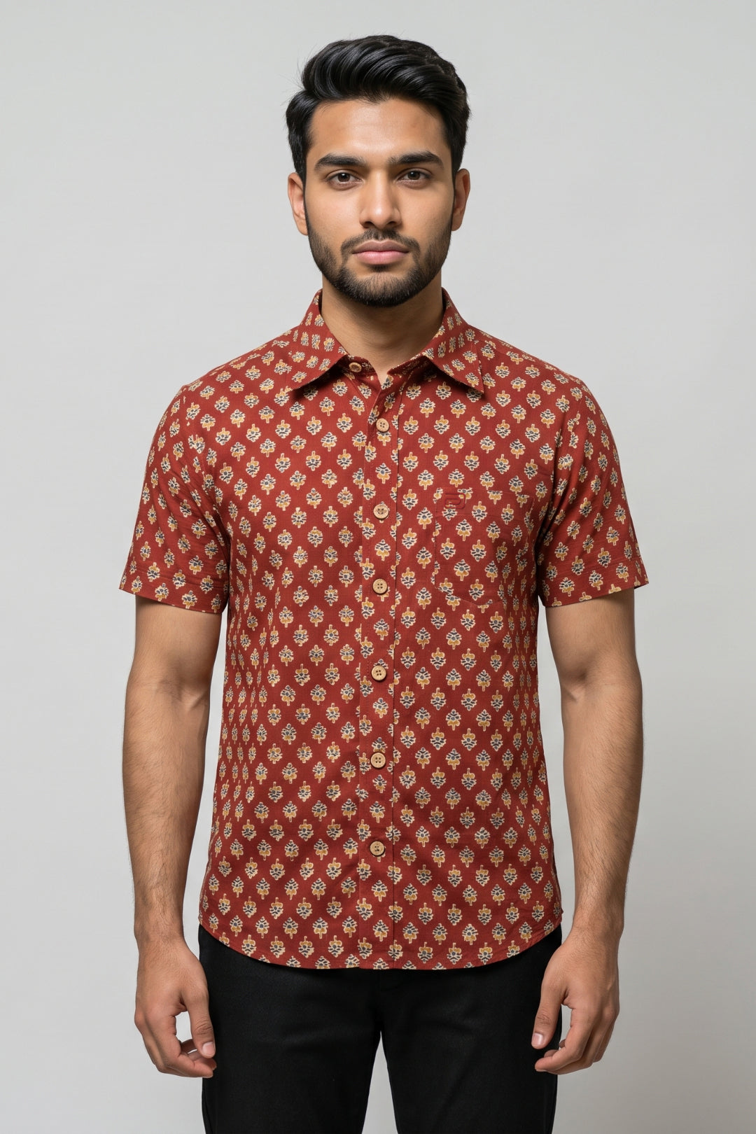 A man wearing a short-sleeve button-up shirt with a rust red and beige geometric pattern, paired with black trousers, standing against a solid gray background.