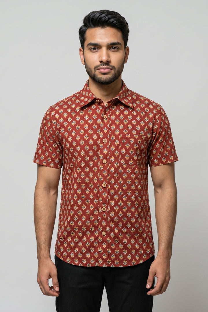 A man wearing a short-sleeve button-up shirt with a rust red and beige geometric pattern, paired with black trousers, standing against a solid gray background.