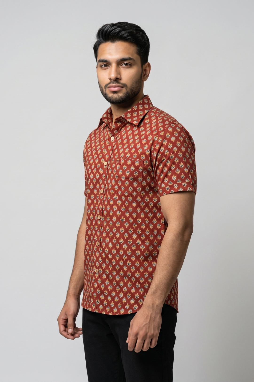 A man wearing a short-sleeve button-up shirt with a red and beige geometric pattern, paired with black pants, standing against a light gray studio background.