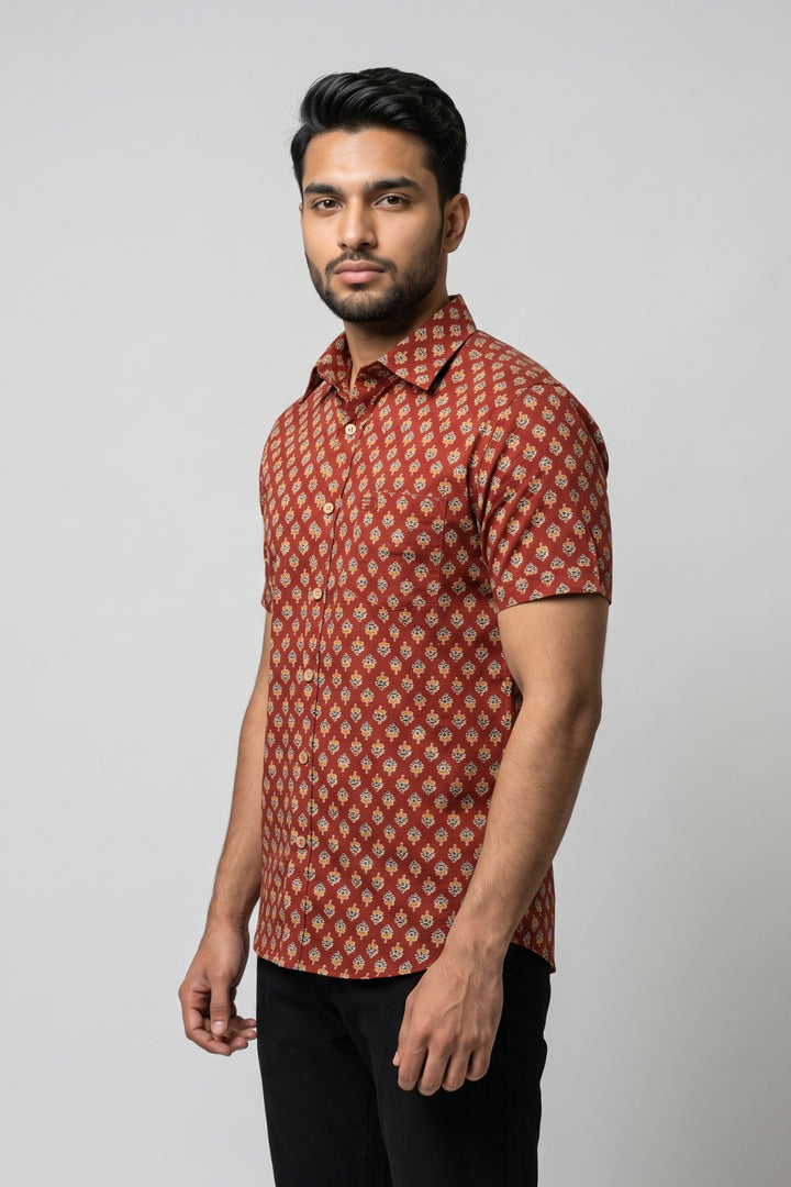 A man wearing a short-sleeve button-up shirt with a red and beige geometric pattern, paired with black pants, standing against a light gray studio background.