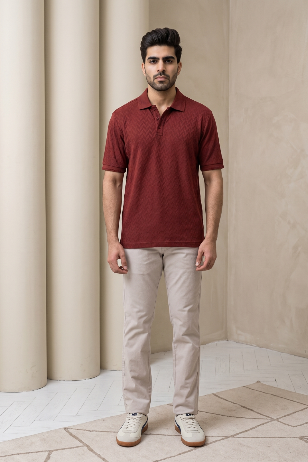 A man wearing a textured maroon short-sleeve polo shirt, beige trousers, and white sneakers, standing in front of a light gray background with columns.