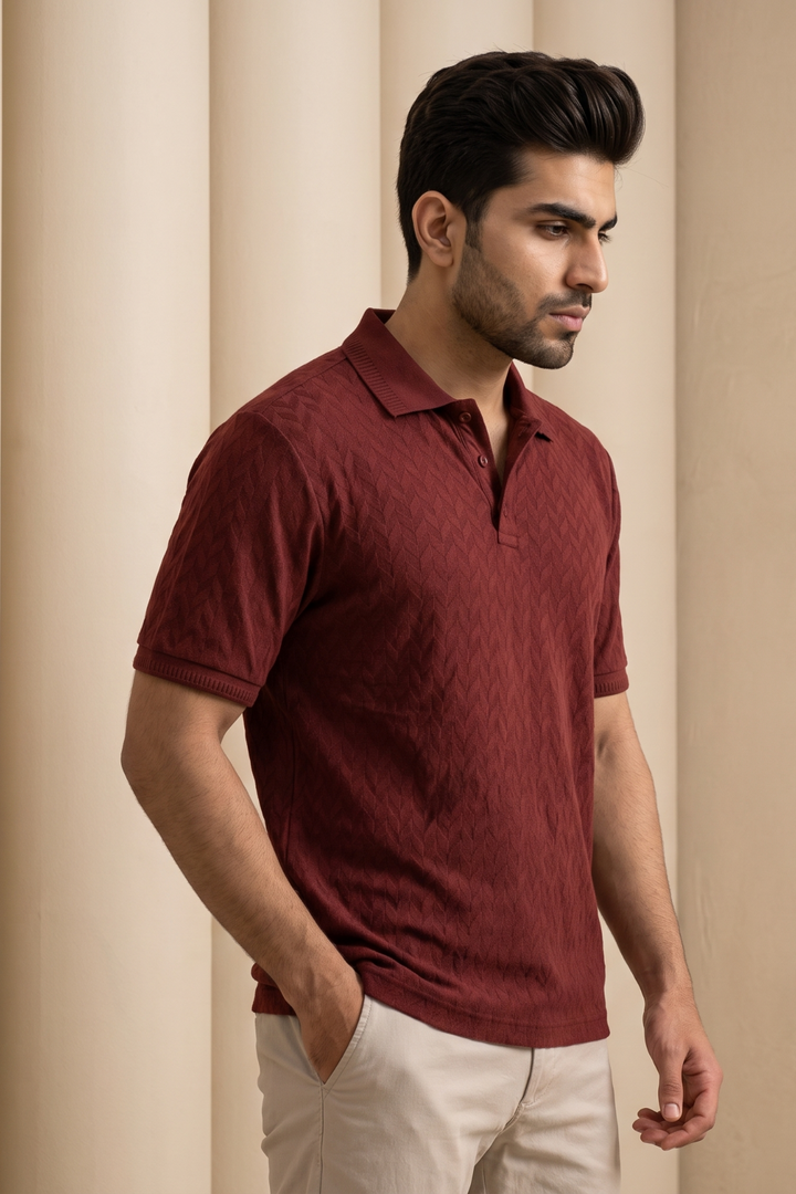 A man wearing a textured maroon short-sleeve polo shirt and light gray trousers, standing against a neutral beige background with soft vertical drapery.