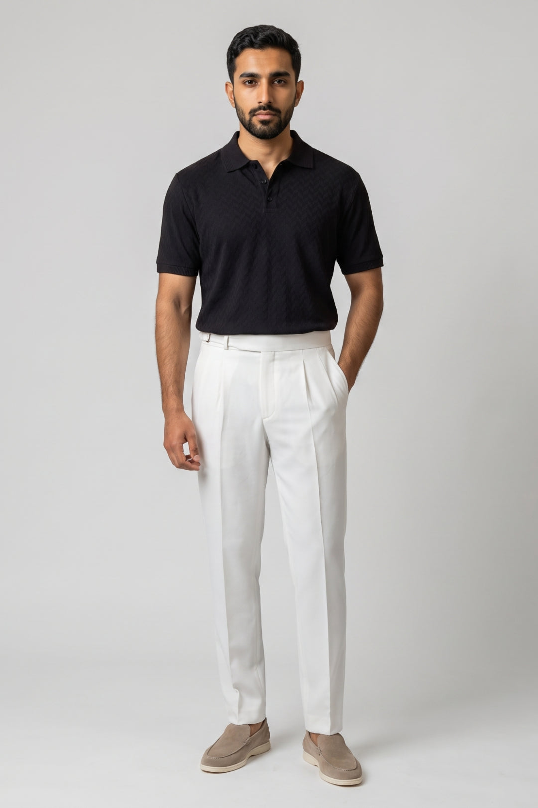 A man modeling a black short-sleeve polo shirt and white trousers with beige slip-on shoes, standing against a light gray studio background.