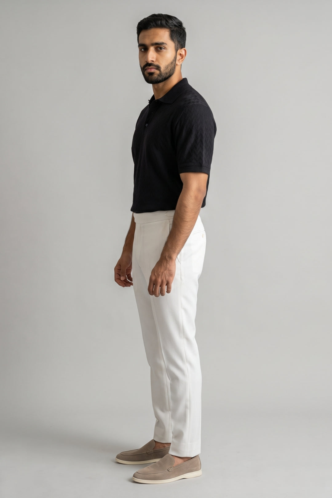 A man standing in profile wearing a black short-sleeve polo shirt, off-white trousers, and beige slip-on shoes against a plain light gray studio background.