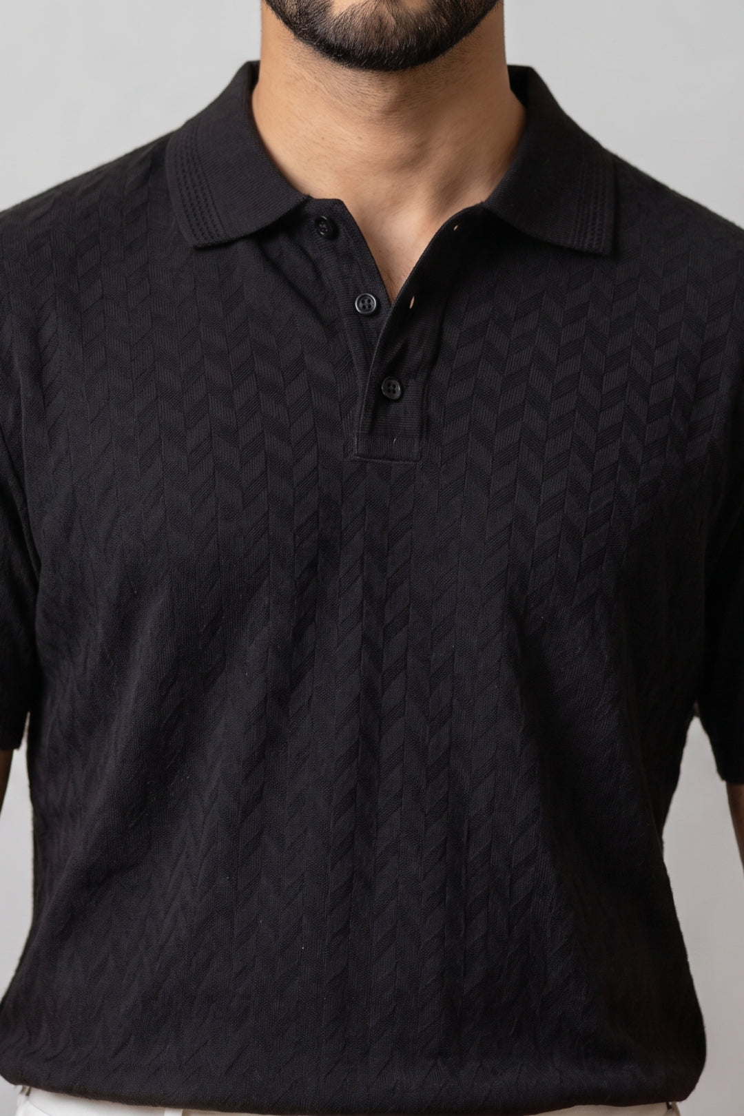 A close-up of a man wearing a black short-sleeve polo shirt with a subtle textured geometric pattern, shown against a neutral gray background.