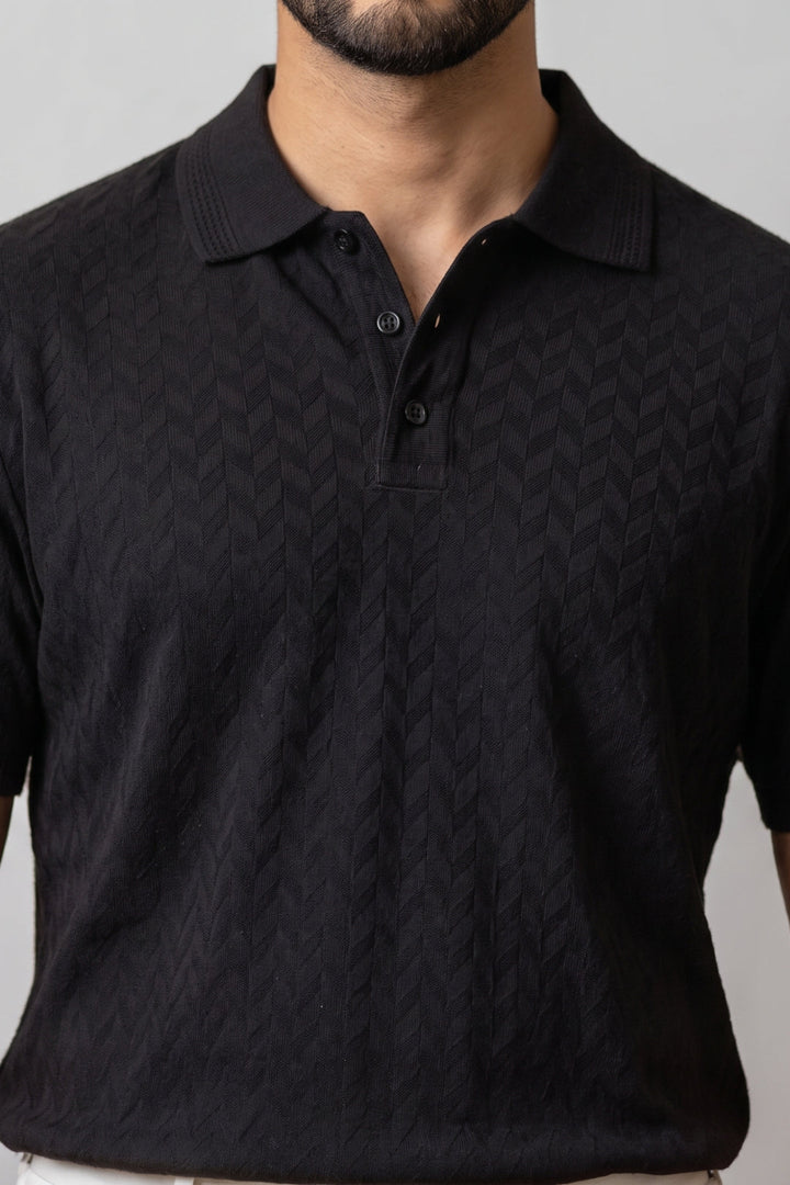 A close-up of a man wearing a black short-sleeve polo shirt with a subtle textured geometric pattern, shown against a neutral gray background.