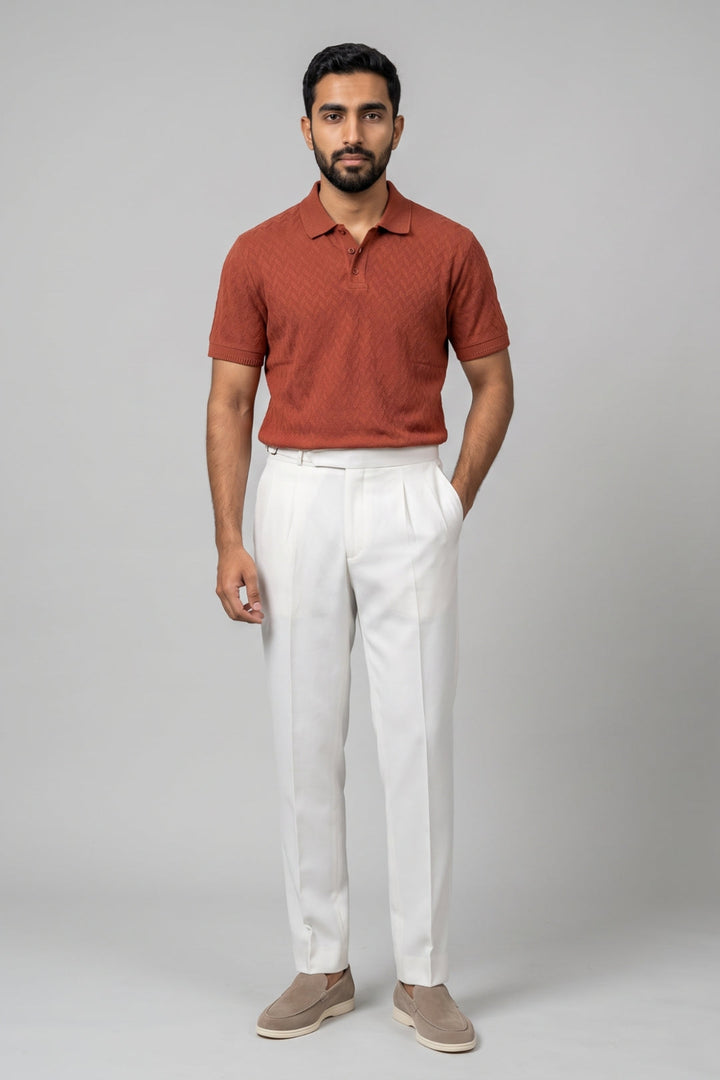 A man modeling a short-sleeve polo shirt in rust orange with a subtle textured pattern, paired with white trousers and beige slip-on shoes, against a solid gray studio background.