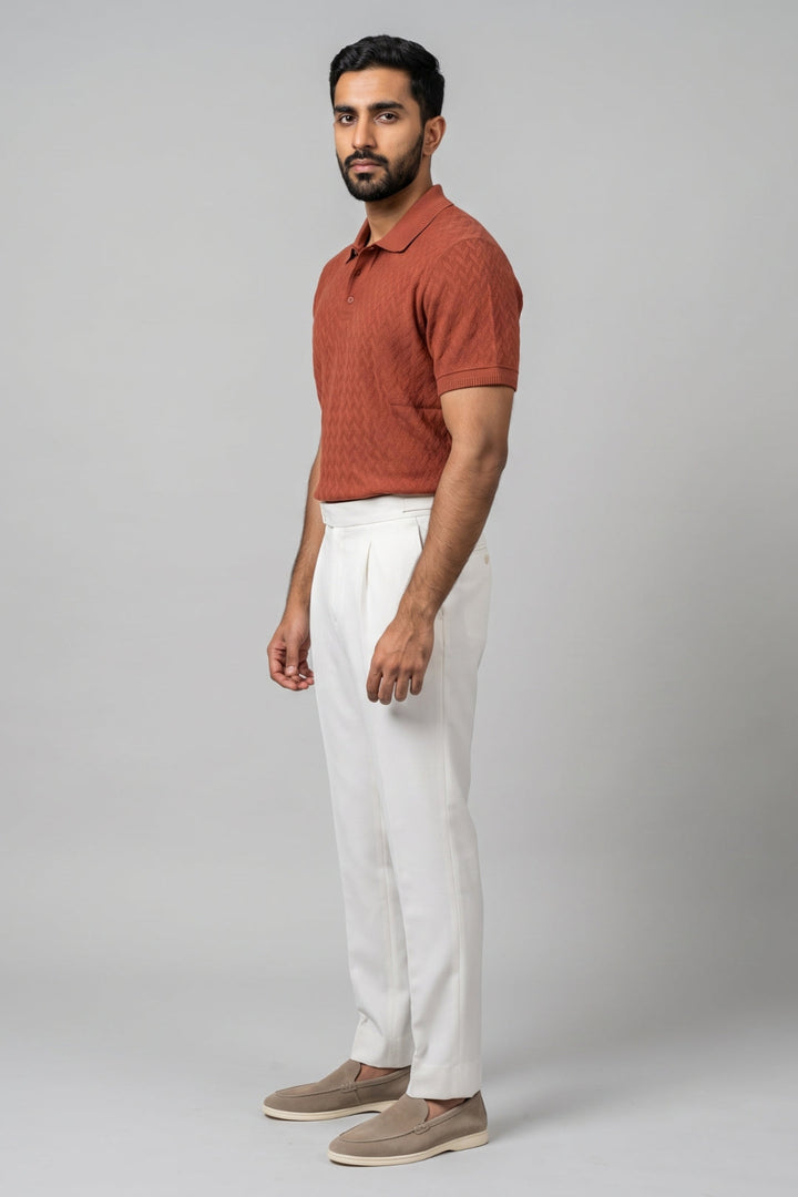A man wearing a rust-orange short-sleeve knit polo shirt, white trousers, and beige slip-on shoes, standing in profile against a light gray studio background.