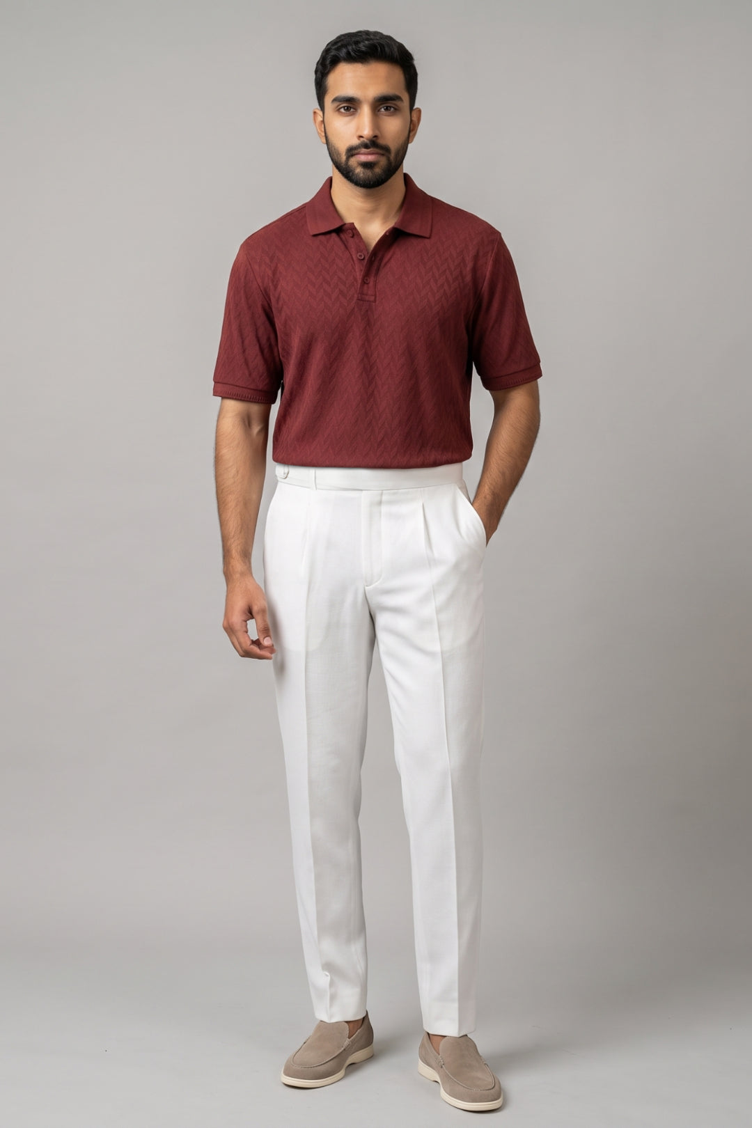 A man wearing a short-sleeve maroon polo shirt with a subtle textured pattern, paired with white trousers and beige slip-on shoes, standing against a solid gray studio background.