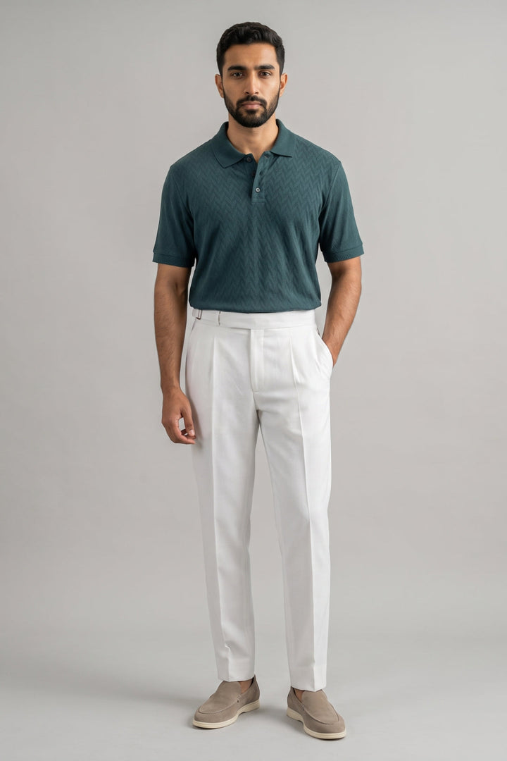 A man modeling a short-sleeve polo shirt in dark teal with a subtle diamond pattern, paired with white pleated trousers and beige slip-on shoes, against a solid light gray background.