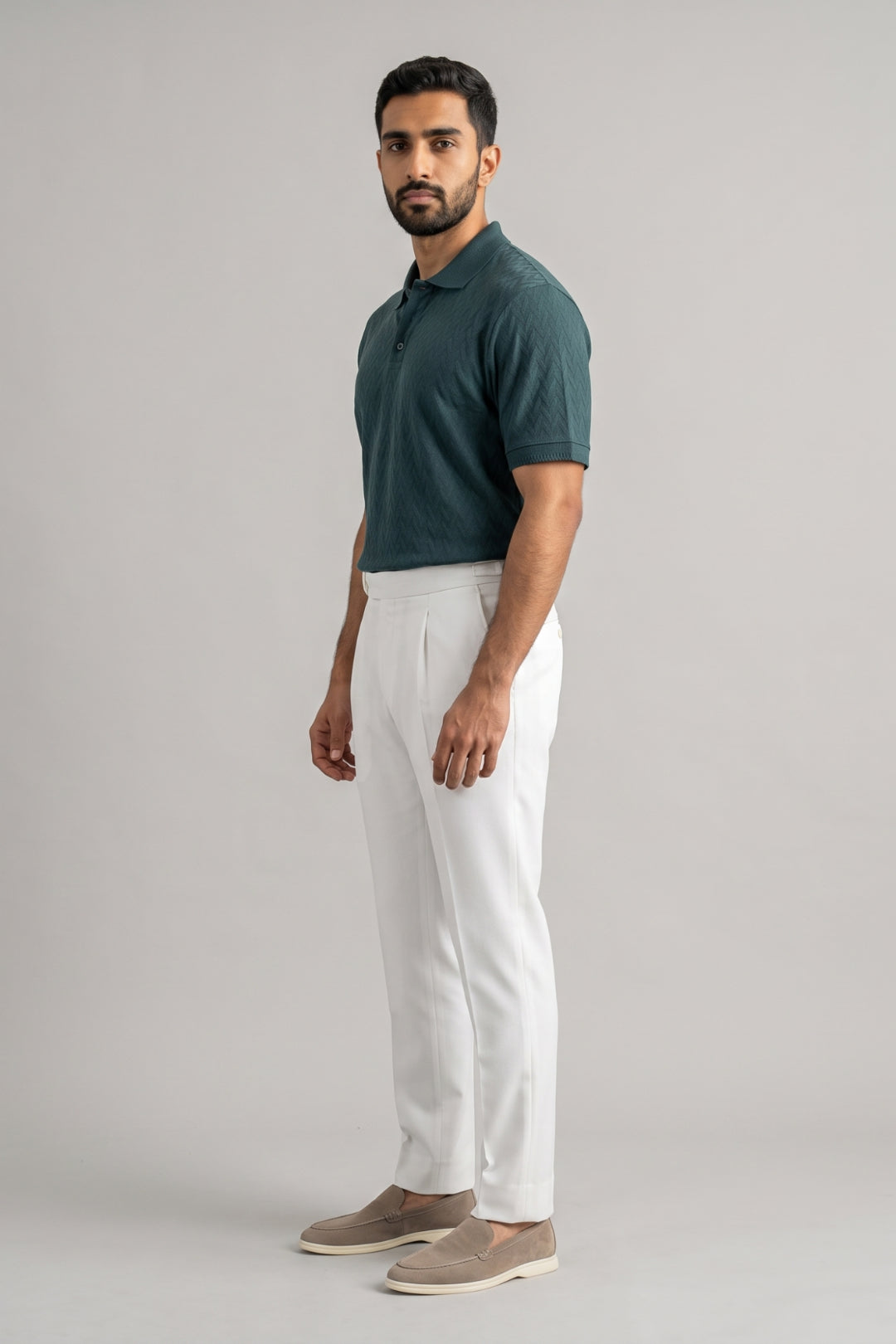 A man modeling a dark green short-sleeve polo shirt and white trousers, standing against a neutral light gray studio background.