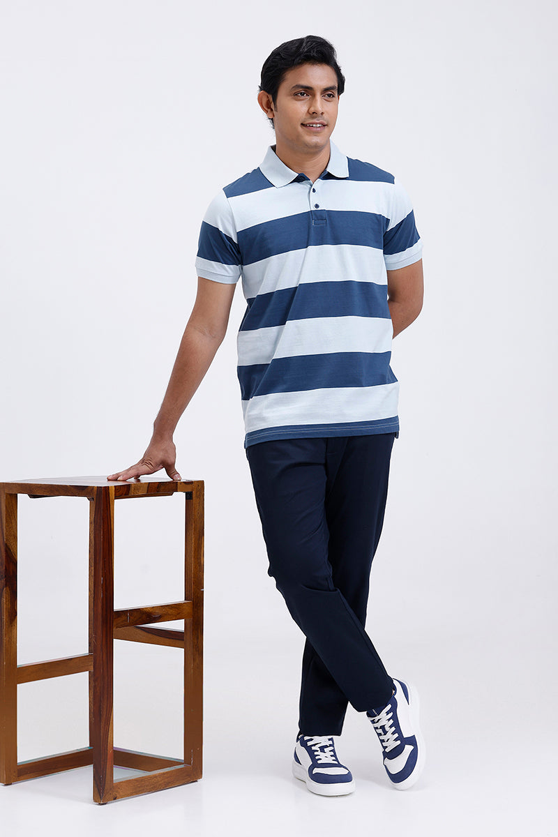 Man wearing a blue and steel blue striped polo shirt with navy pants, standing next to a wooden stool on a white background.
