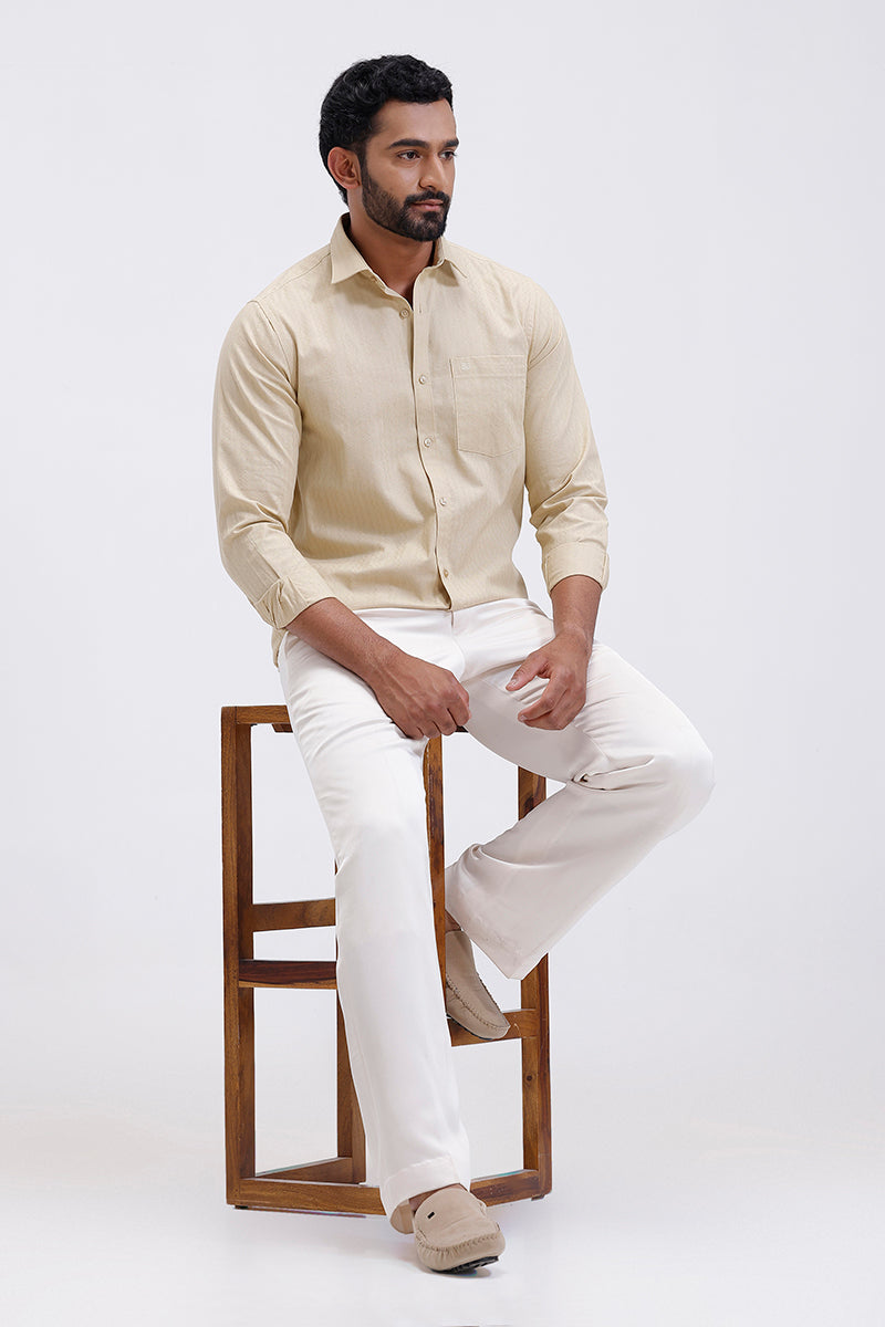 Man sitting on a wooden stool wearing a beige shirt and white pants against a white background