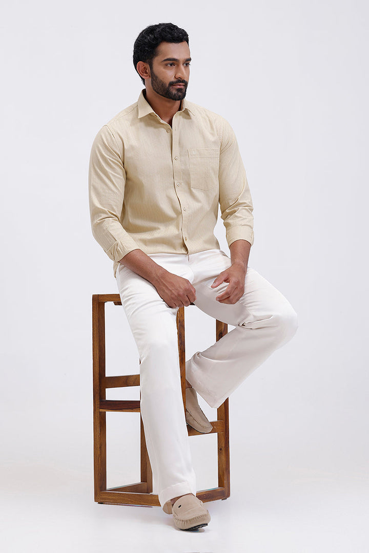 Man sitting on a wooden stool wearing a beige shirt and white pants against a white background