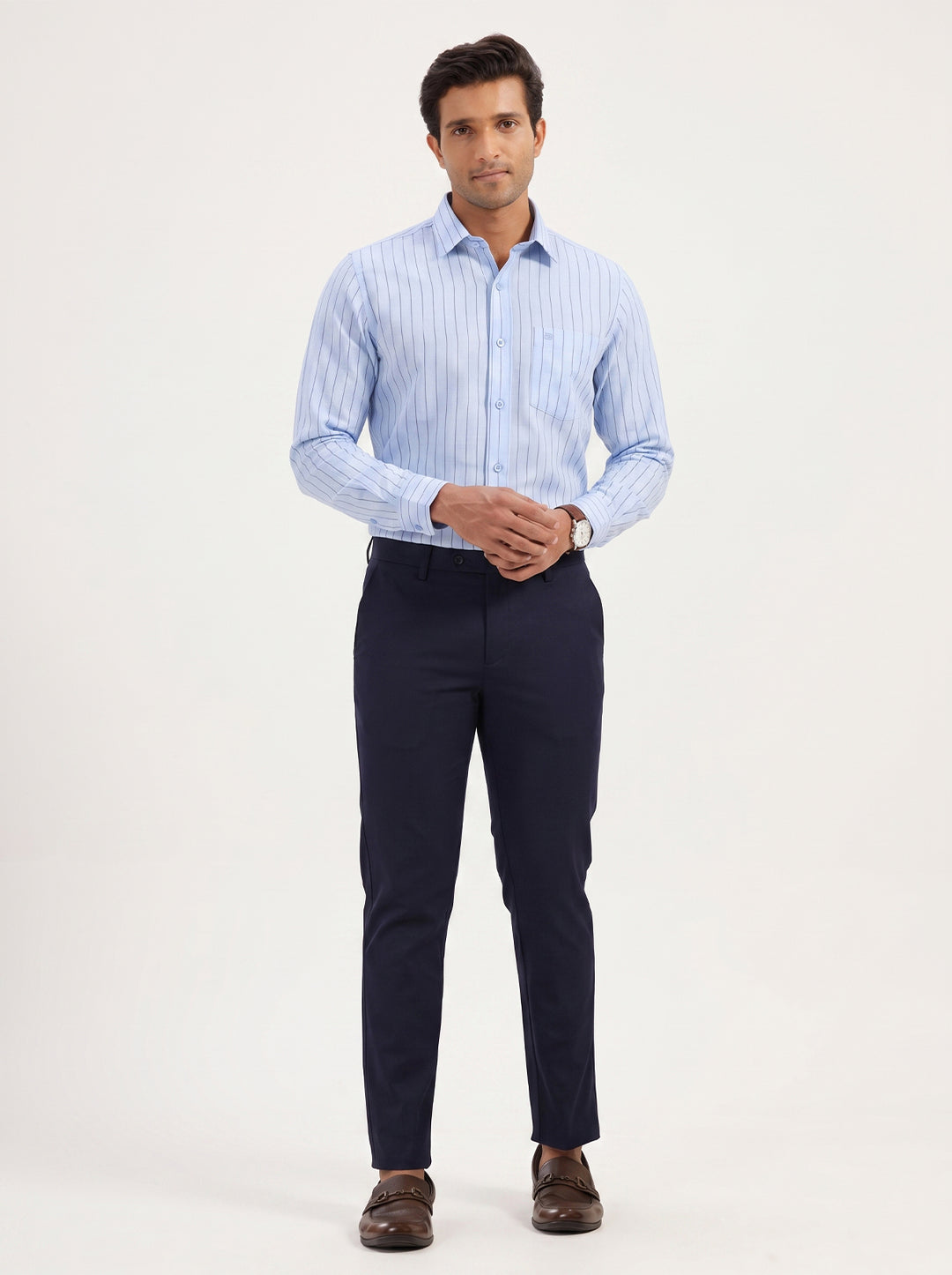 A man modeling a light blue and white striped long-sleeve dress shirt paired with navy blue trousers and brown leather dress shoes, standing against a plain off-white studio background.