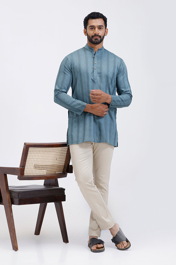 Man wearing a steel blue kurta with beige pants standing next to a wooden chair on a white background