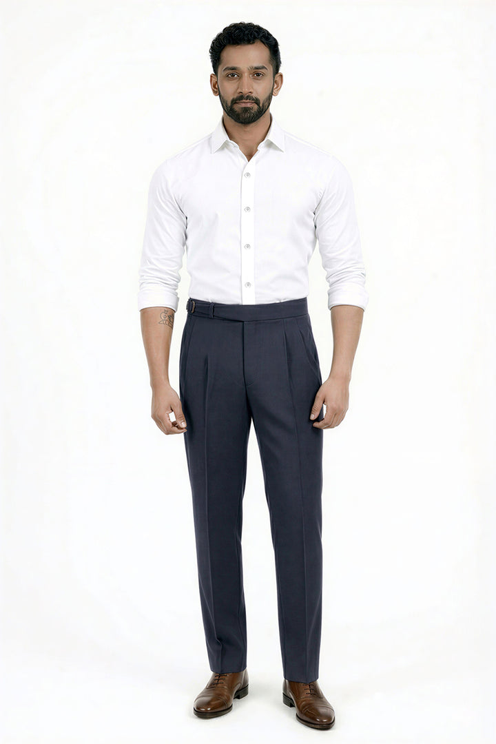 A man modeling a white long-sleeve button-down shirt and dark navy blue pleated trousers with brown leather dress shoes, standing against a plain white studio background.
