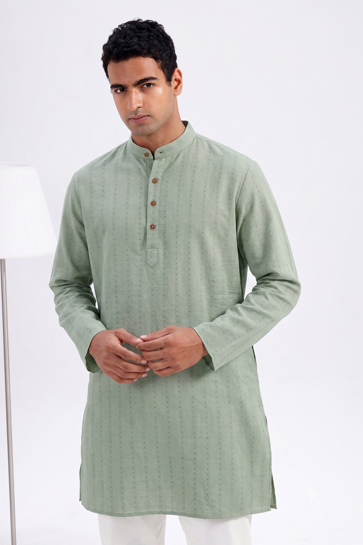 A man wearing a green kurta with a mandarin collar and three brown buttons, paired with white trousers, standing against a white studio background.