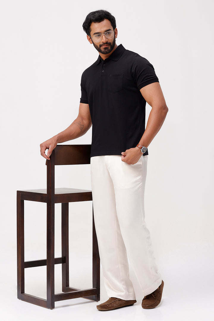 Man wearing a deep black polo shirt and white pants standing next to a wooden chair on a white background