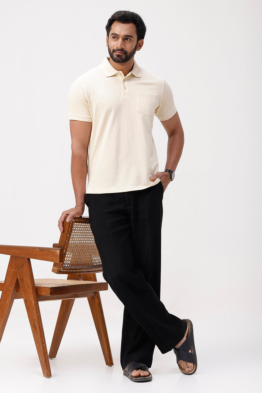 Man wearing a white polo shirt and black pants standing next to a wooden chair on a white background
