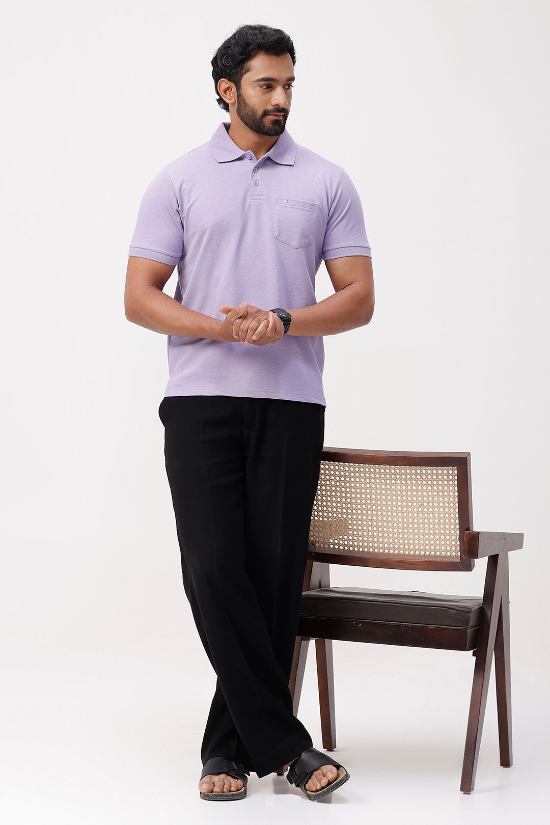 Man wearing a soft lavender polo shirt and black pants standing next to a wooden chair on a white background