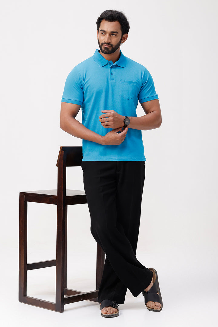 Man wearing a ice blue polo shirt and black pants standing next to a wooden chair on a white background