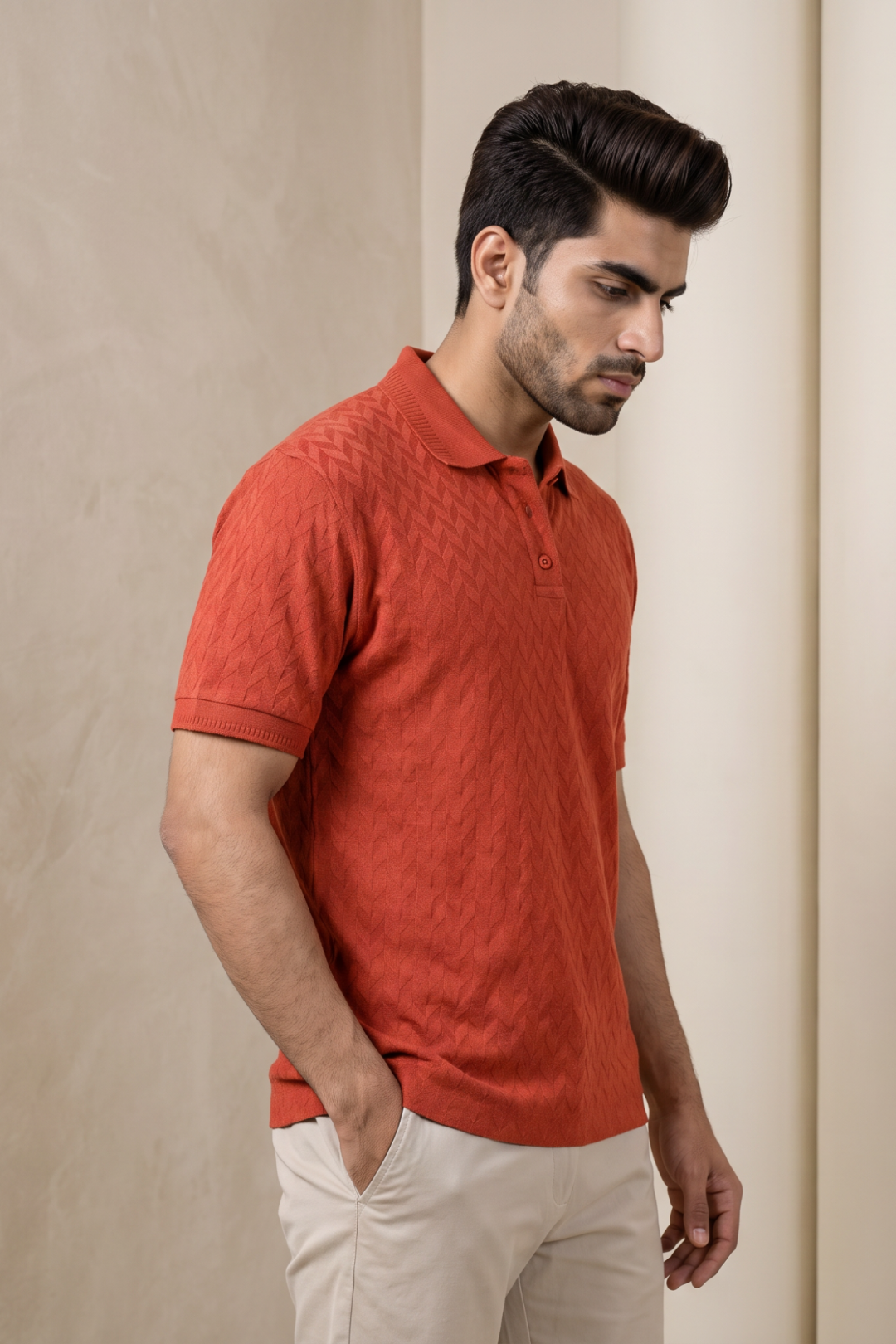 A man wearing a textured rust-orange short-sleeve polo shirt and light beige trousers, standing against a neutral beige wall.