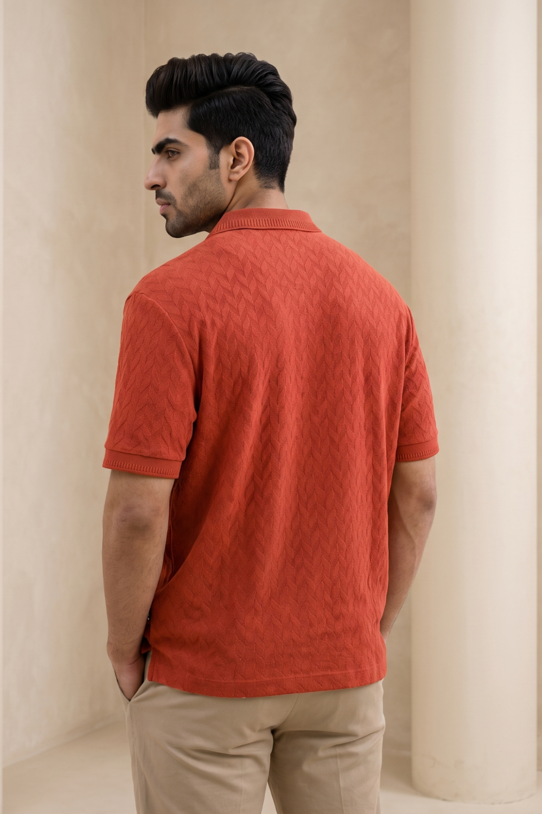 A man wearing a textured terra orange short-sleeve polo shirt, shown from the back in a warm, neutral indoor setting with beige columns.