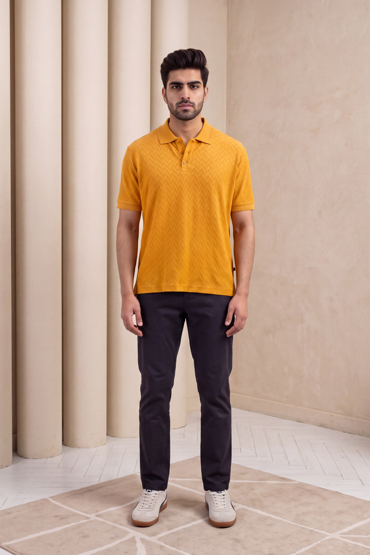A man stands facing forward wearing a mustard yellow textured short-sleeve polo shirt, dark navy trousers, and white sneakers in a minimalist beige interior setting with columns.